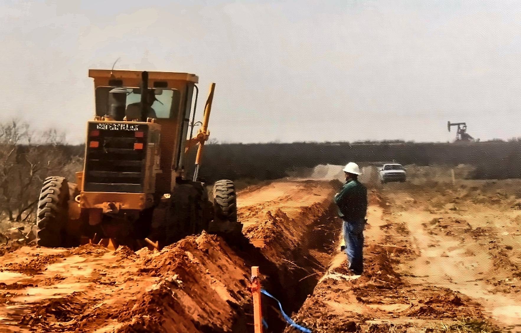 Motor grader preparing a pipeline right-of-way with a pump jack in the distance